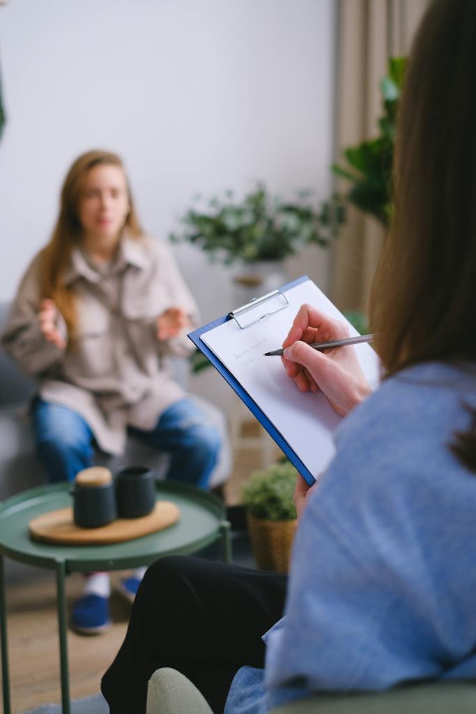 Unrecognizable female psychologist taking notes on clipboard while listening to patient sitting on blurred background during psychotherapy consultation in office